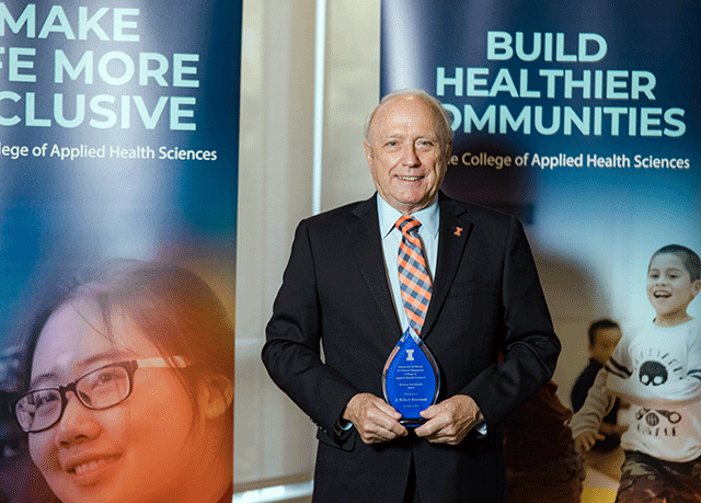 Dr. J. Robert Rossman holding award and standing in front of banners