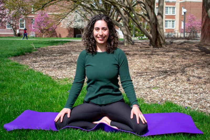 A woman in a green sweater sits in the grass with a purple yoga mat. 