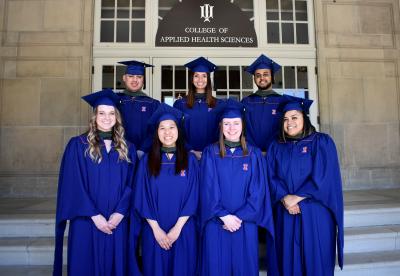 7 students in graduation gowns posing in front of Huff Hall